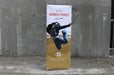 Eco-friendly bamboo x-stand banner display featuring custom printed graphic "WE PRINT BANNER STANDS ask us how!" set outdoors against a concrete wall.