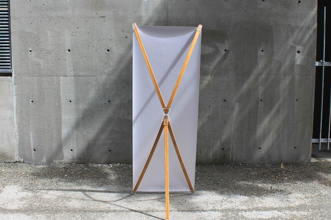Rear view of a bamboo X-stand banner display showing the X-frame bamboo leg structure, center hub, and back support leg against a concrete wall.