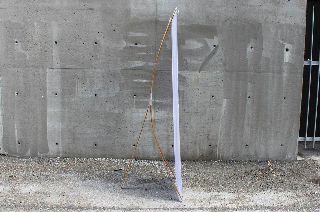 Side profile view of a bamboo X-stand display showing the flexible bamboo support arms hooked to the vertical white banner against a concrete wall.