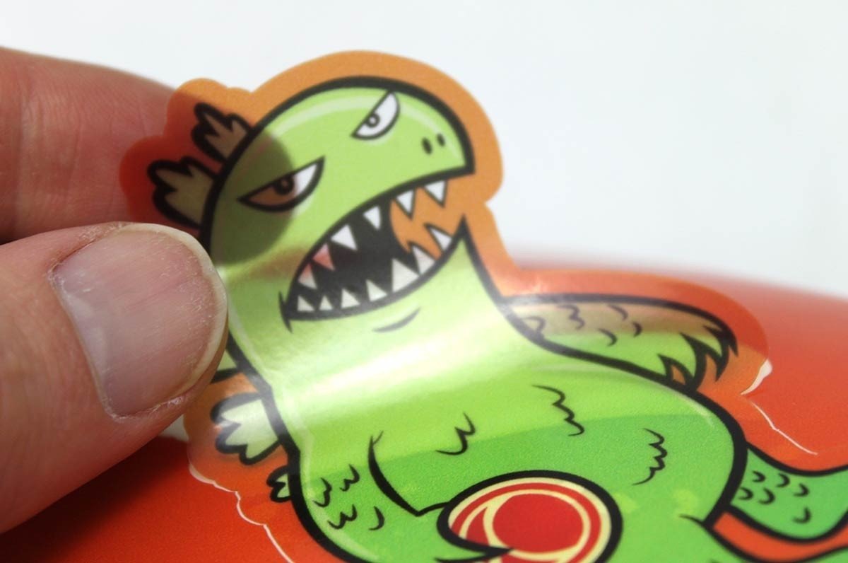 Hand peeling a clear custom shape vinyl sticker sample featuring a Clubcard's green cartoon dinosaur mascot illustration, printed by Clubcard Vancouver