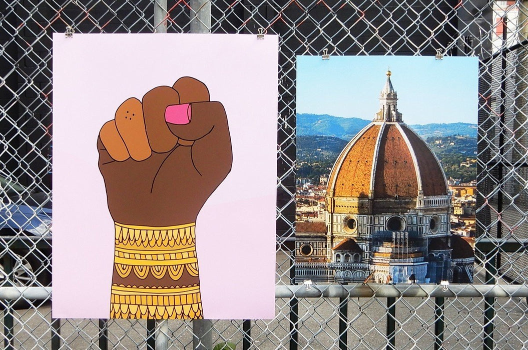 Two large fine art print samples displayed on a chain-link fence, featuring an artwork of a raised fist by artist Jag Nagra with gold bangles and a photo of the Florence Duomo, printed by Clubcard Vancouver