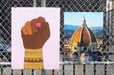 Two large fine art print samples displayed on a chain-link fence, featuring an artwork of a raised fist by artist Jag Nagra with gold bangles and a photo of the Florence Duomo, printed by Clubcard Vancouver