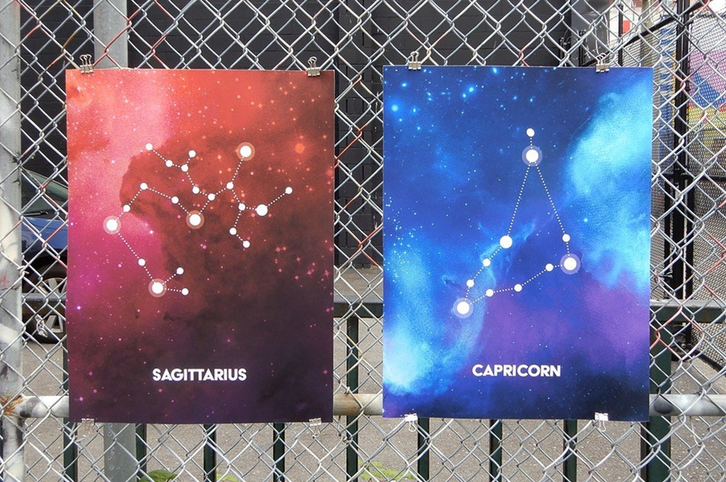 Two large fine art print samples displayed on a chain-link fence featuring Sagittarius and Capricorn zodiac constellation illustrations in vibrant galaxy art, printed by Clubcard