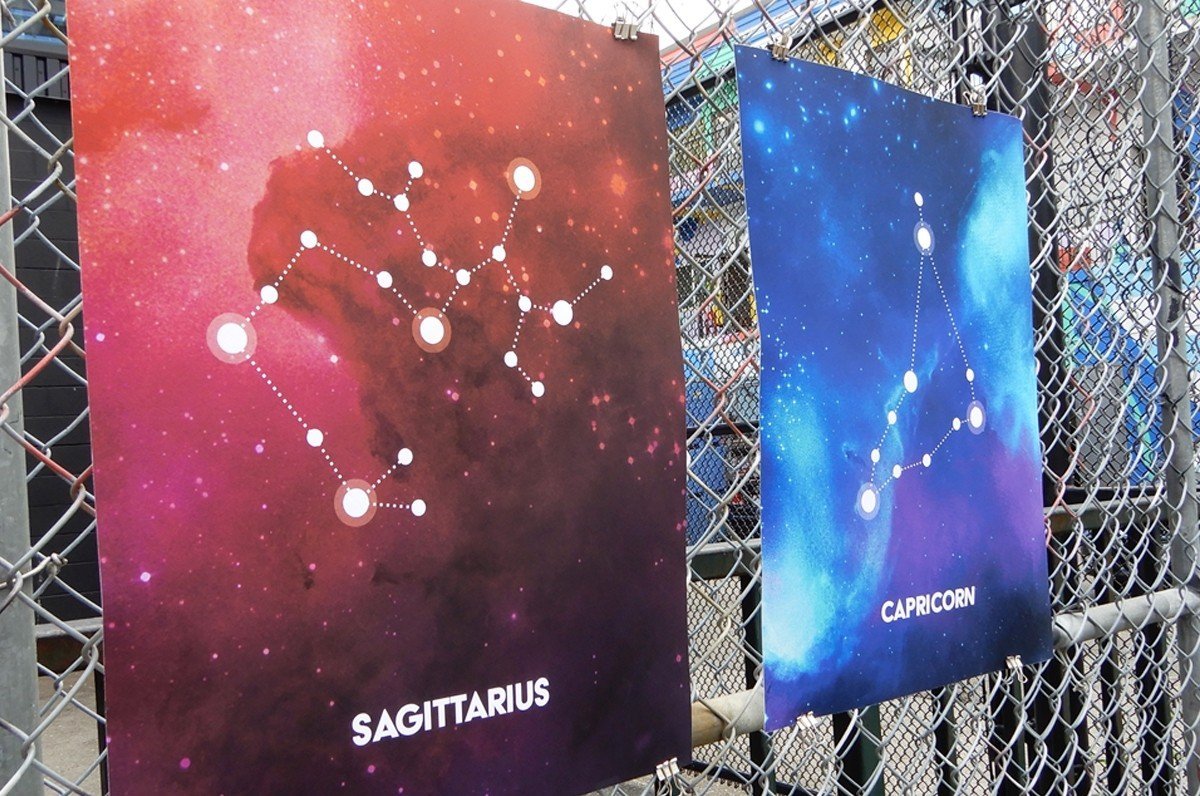 Two large fine art print samples displayed on a chain-link fence featuring Sagittarius and Capricorn zodiac constellation illustrations in vibrant galaxy art, printed by Clubcard