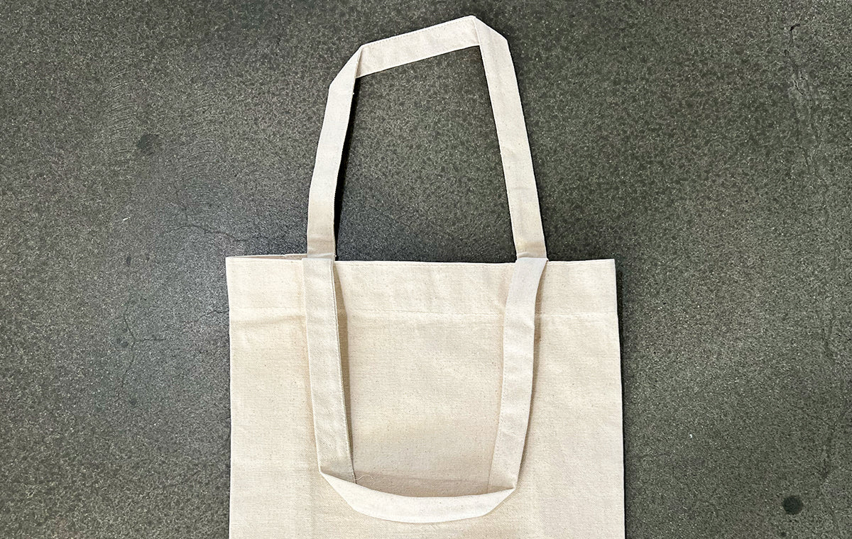 Plain natural canvas tote bag sample lying flat on a dark concrete background, printed by Clubcard Vancouver