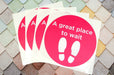 Fanned-out stack of custom white square vinyl sheets printed with bright pink oval floor stickers reading "A great place to wait" with footprint icons.
