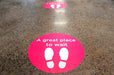 Bright pink oval vinyl floor sticker printed with "A great place to wait" and footprints icon applied to a speckled terrazzo floor