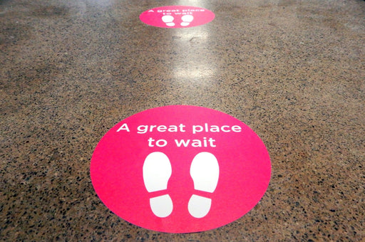 Bright pink oval vinyl floor sticker printed with "A great place to wait" and footprints icon applied to a speckled terrazzo floor