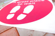 A fanned-out stack of bright pink circular vinyl floor stickers printed with white footprints and "A great place to wait" text, presented on white square backing sheets against a tile background.