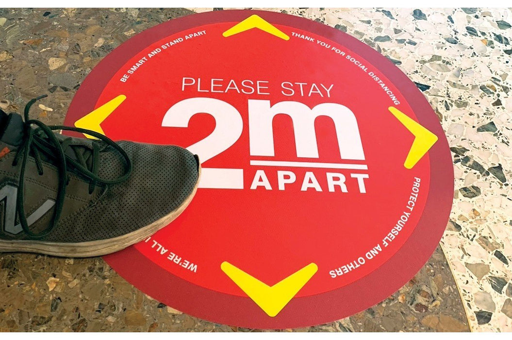 A shoe stepping near a round red and yellow floor decal reading Please Stay 2m Apart for social distancing.
