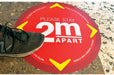 A shoe stepping near a round red and yellow floor decal reading Please Stay 2m Apart for social distancing.