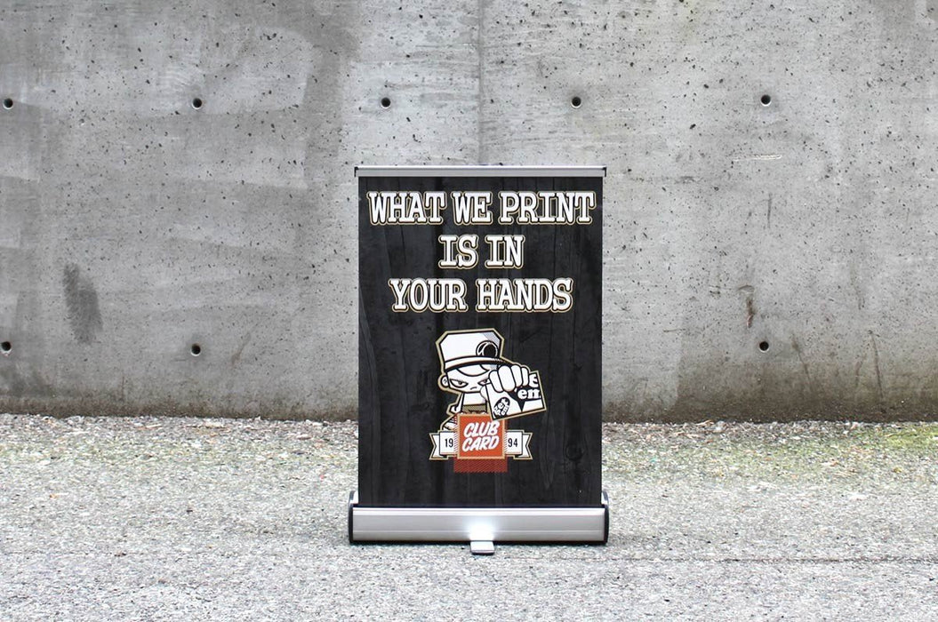 Extra large 18x24.5 inch table-top retractable banner stand displaying custom artwork "What We Print Is In Your Hands" with a silver base against a concrete wall.