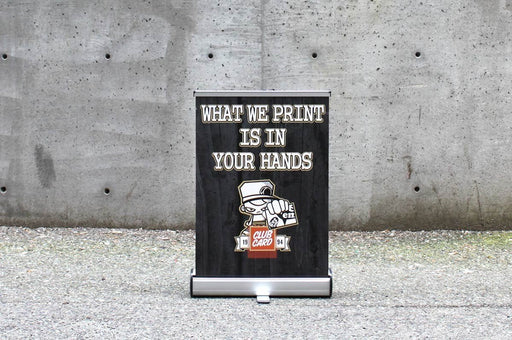 Extra large 18x24.5 inch table-top retractable banner stand displaying custom artwork "What We Print Is In Your Hands" with a silver base against a concrete wall.