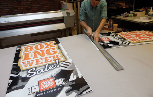 Print shop technician using a ruler and utility knife to trim printed Boxing Week Sale replacement banners