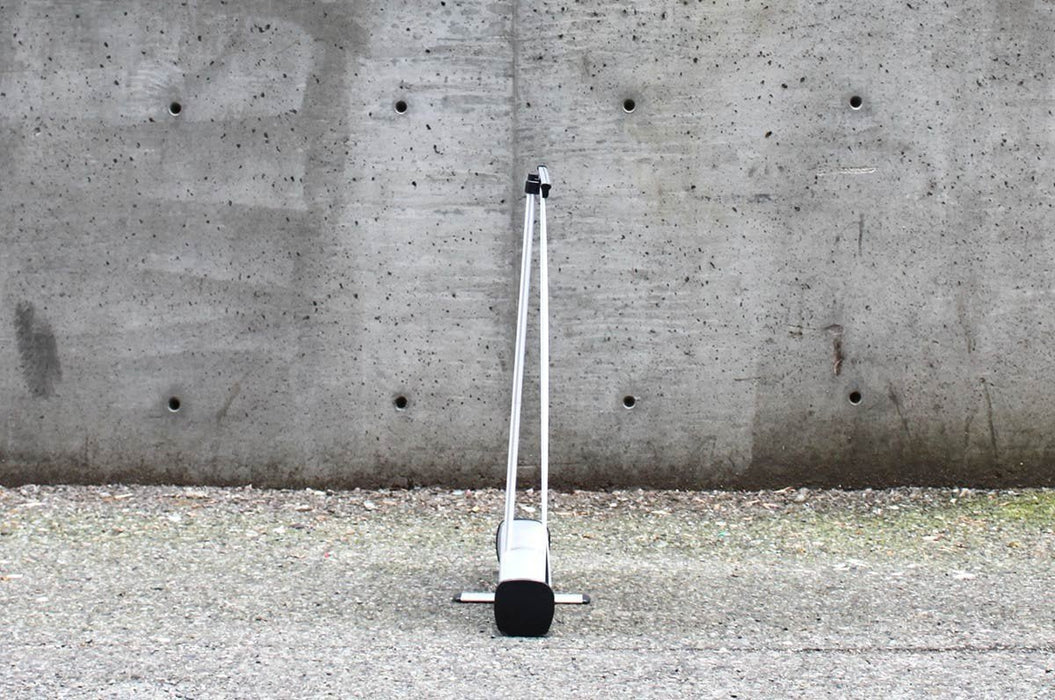 Rear view of an extra large table-top retractable banner stand displaying the silver aluminum base and vertical support pole set against a concrete wall.