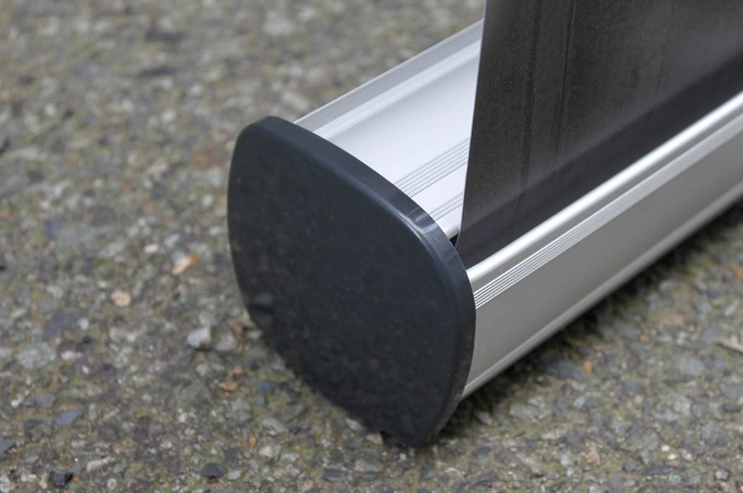 Close-up side view of the silver aluminum cassette base and dark gray plastic end cap of an extra-large tabletop retractable banner stand resting on pavement.