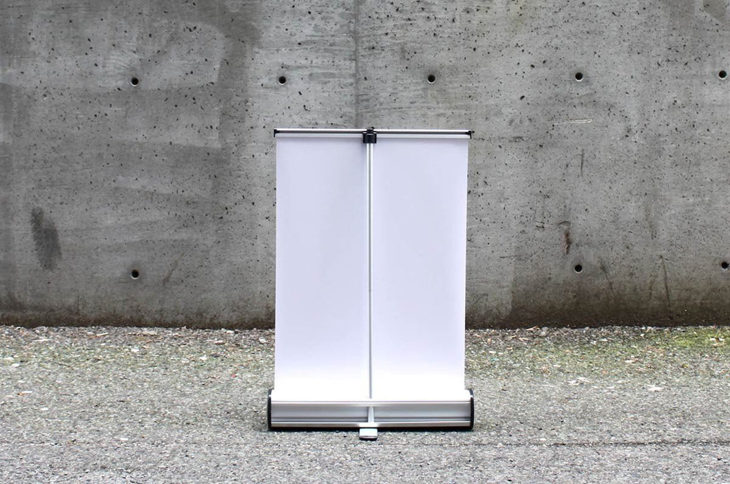 Rear view of a blank white extra-large tabletop retractable banner stand showing the silver base and rear support pole against a concrete wall.