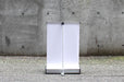 Rear view of a blank white extra-large tabletop retractable banner stand showing the silver base and rear support pole against a concrete wall.