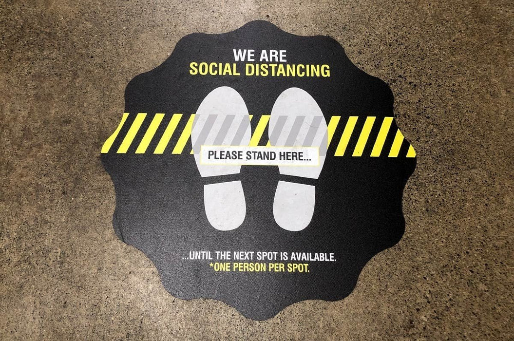 Black social distancing floor decal reading We Are Social Distancing Please Stand Here with footprint graphics on concrete