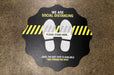 Black social distancing floor decal reading We Are Social Distancing Please Stand Here with footprint graphics on concrete