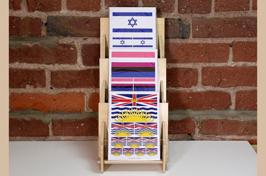 Slim birch plywood 3-tier display card stand holding three types of flag stickers in front of a brick wall.